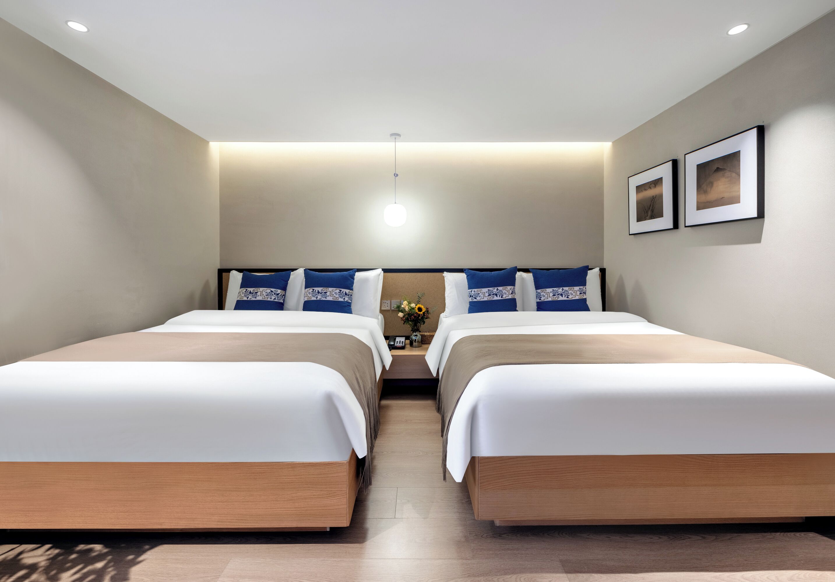 A hotel room with two beds, a nightstand with a vase of flowers, and two framed pictures on the wall.