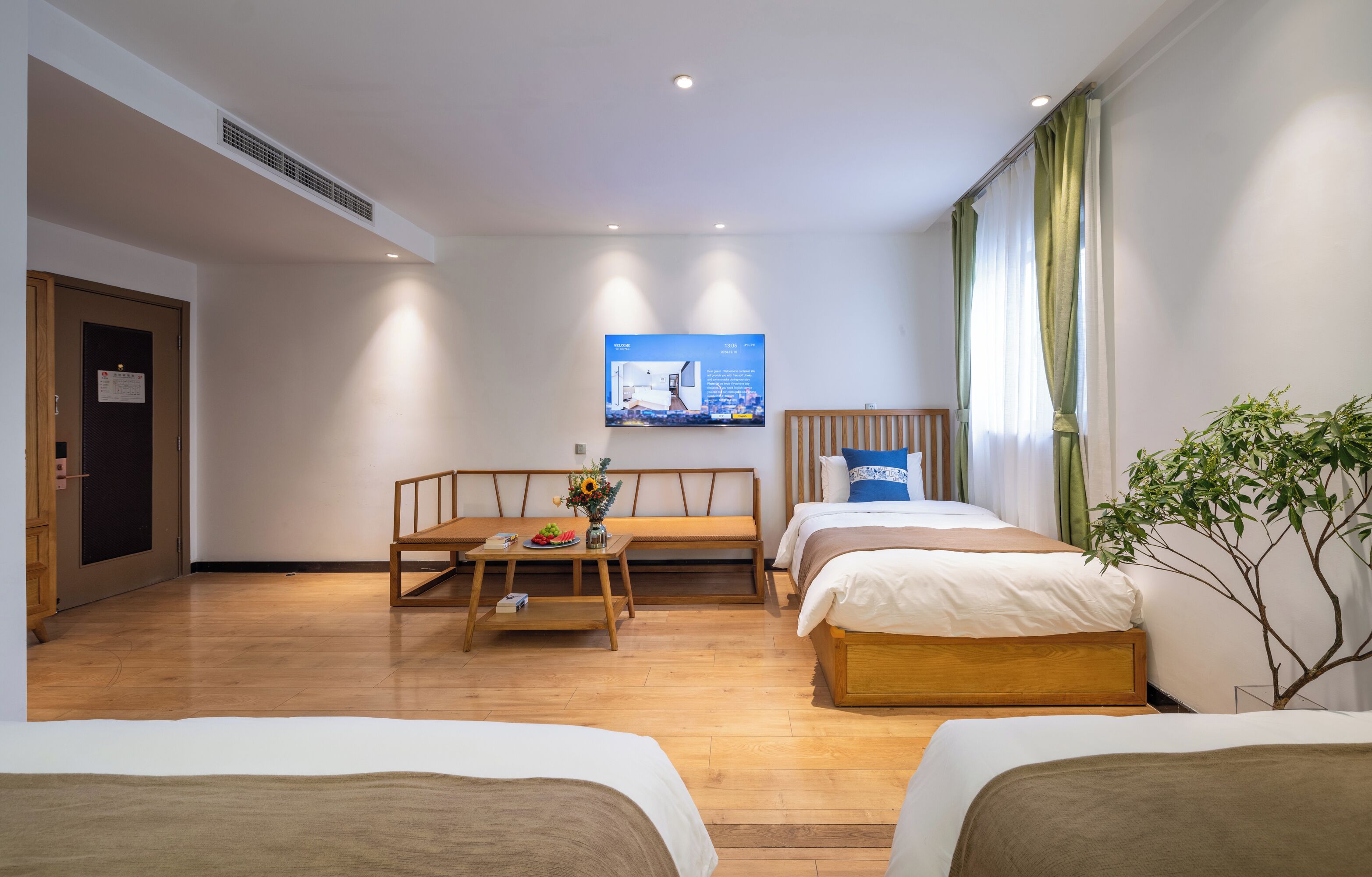 Une chambre d’hôtel avec deux lits, un banc en bois, une petite table avec un vase et des fruits, et un téléviseur fixé au mur.
