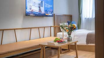 Ein Hotelzimmer mit Bett, einer hölzernen Bank, einem Tisch mit Früchten und einer Vase sowie einem Fernseher, der eine Stadtansicht zeigt.