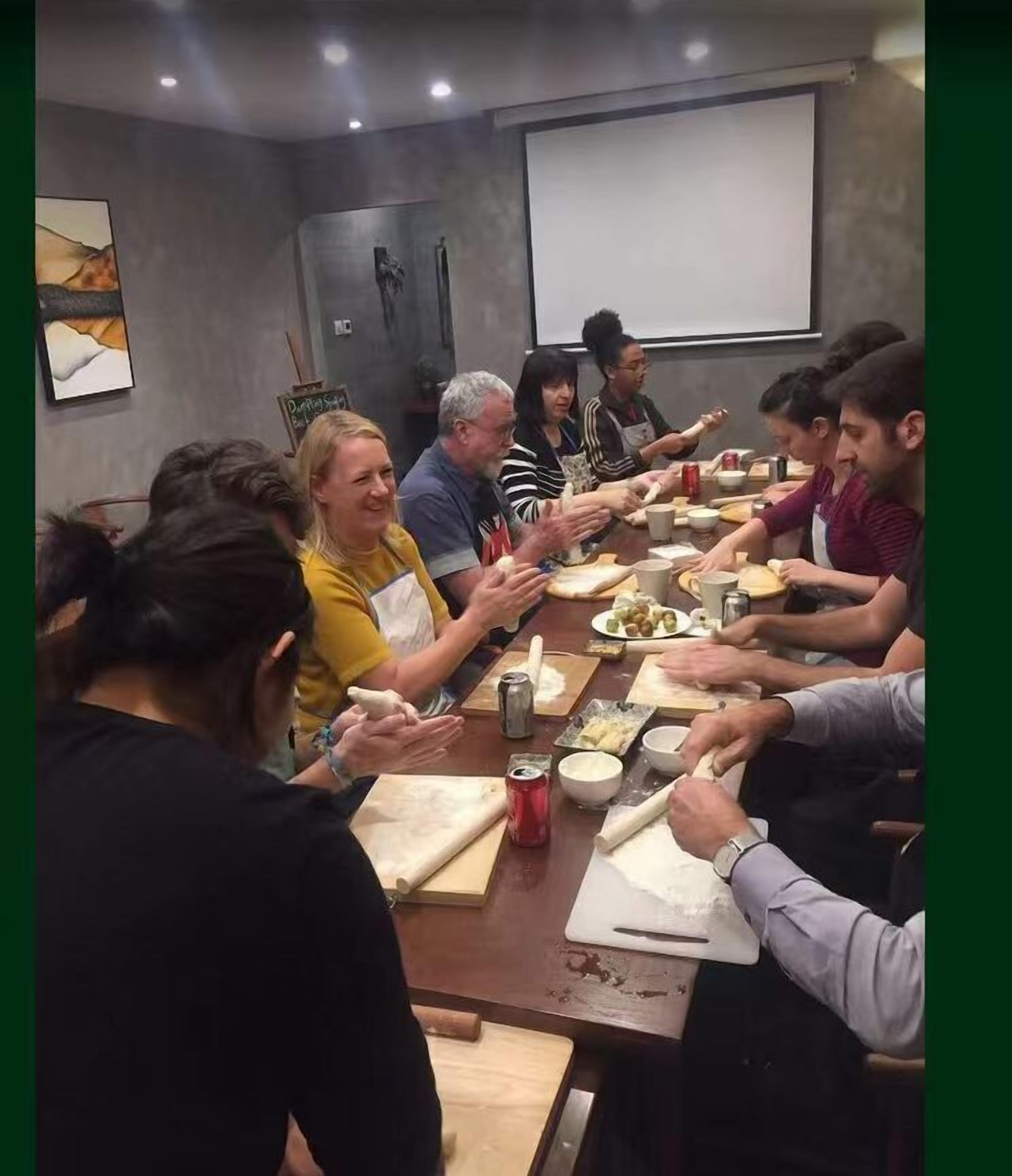 A group of people engaged in a cooking activity around a long table with various cooking utensils and ingredients.