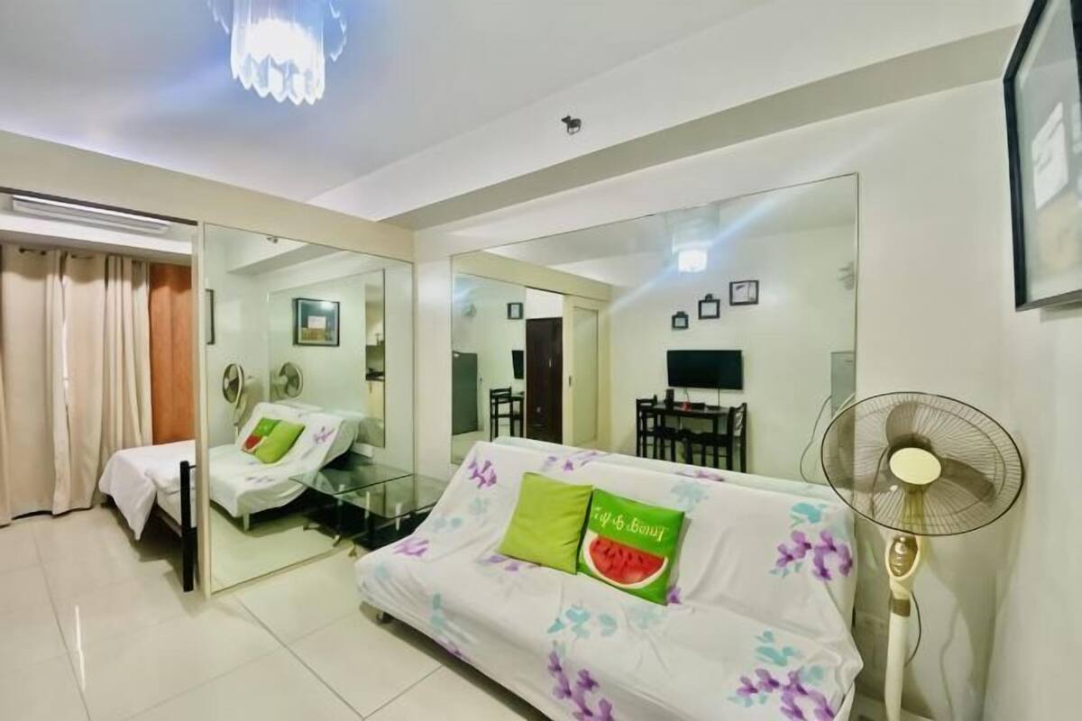 A modern hotel room with a mirrored wall, a sofa with floral upholstery, a standing fan, and a bed with a white headboard.