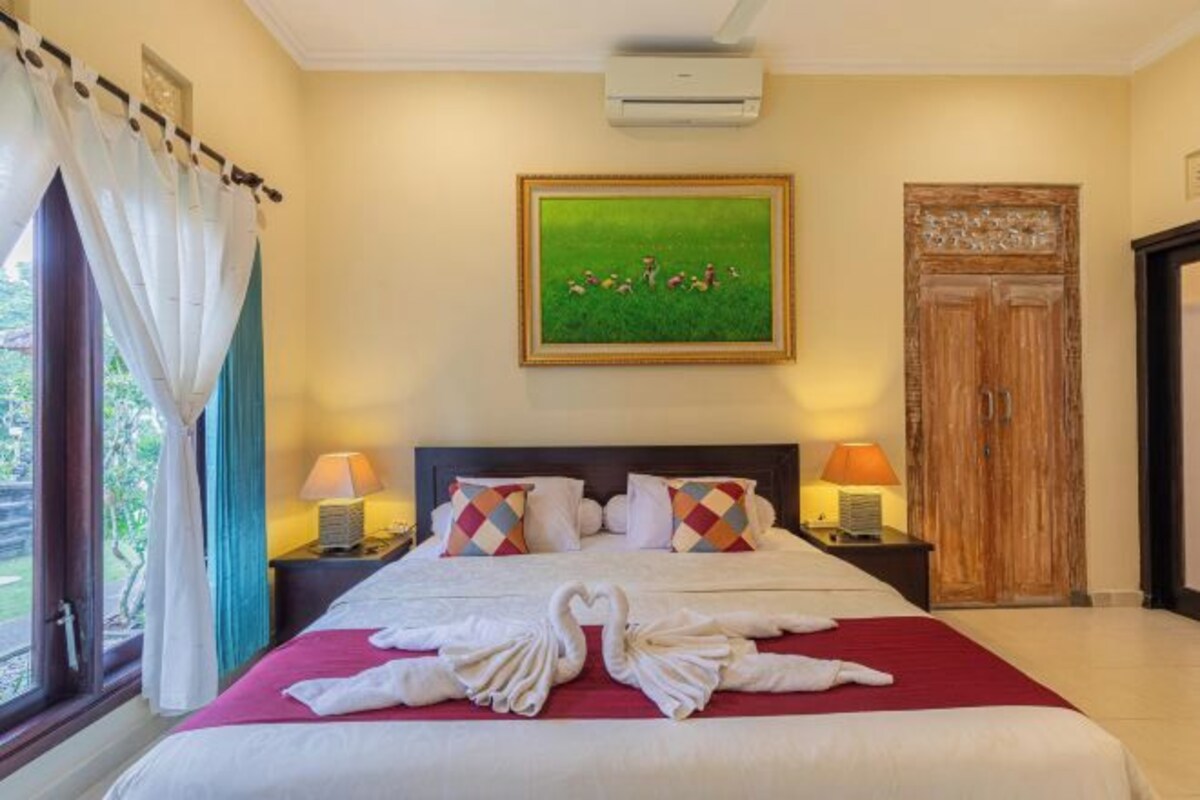 Une chambre d’hôtel avec un lit, deux tables de chevet avec des lampes, un tableau encadré au mur, une porte en bois et une fenêtre avec des rideaux.