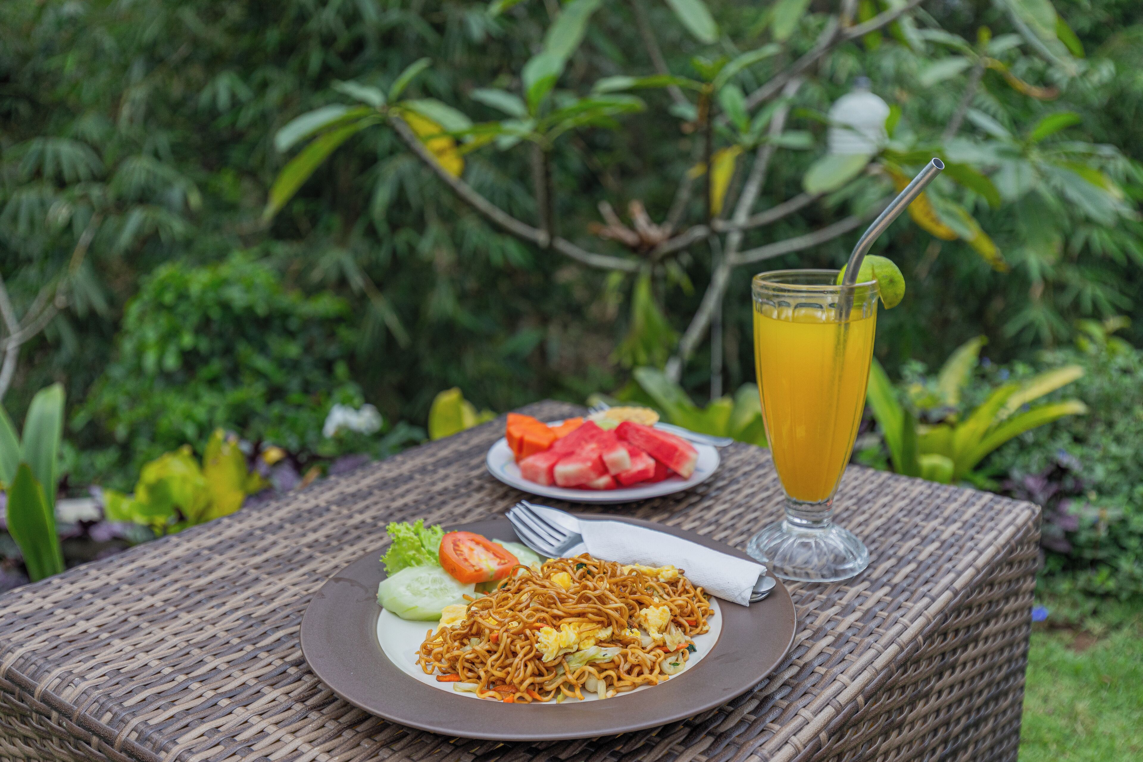A wicker table with a plate of noodles, a glass of orange juice, and a plate of watermelon and tomatoes.