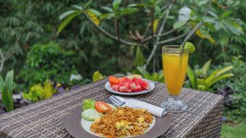 A wicker table with a plate of noodles, a glass of orange juice, and a plate of watermelon and tomatoes.