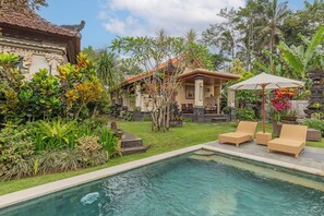 A pool area with lounge chairs and a covered seating area, surrounded by lush tropical plants and a traditional building with a tiled roof.