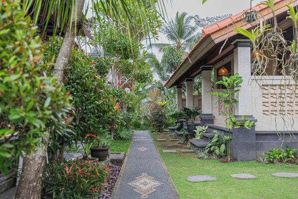 A pathway leading to a building with a tiled roof and columns, surrounded by lush greenery and potted plants. - Campuhan Guesthouse (Penebel)