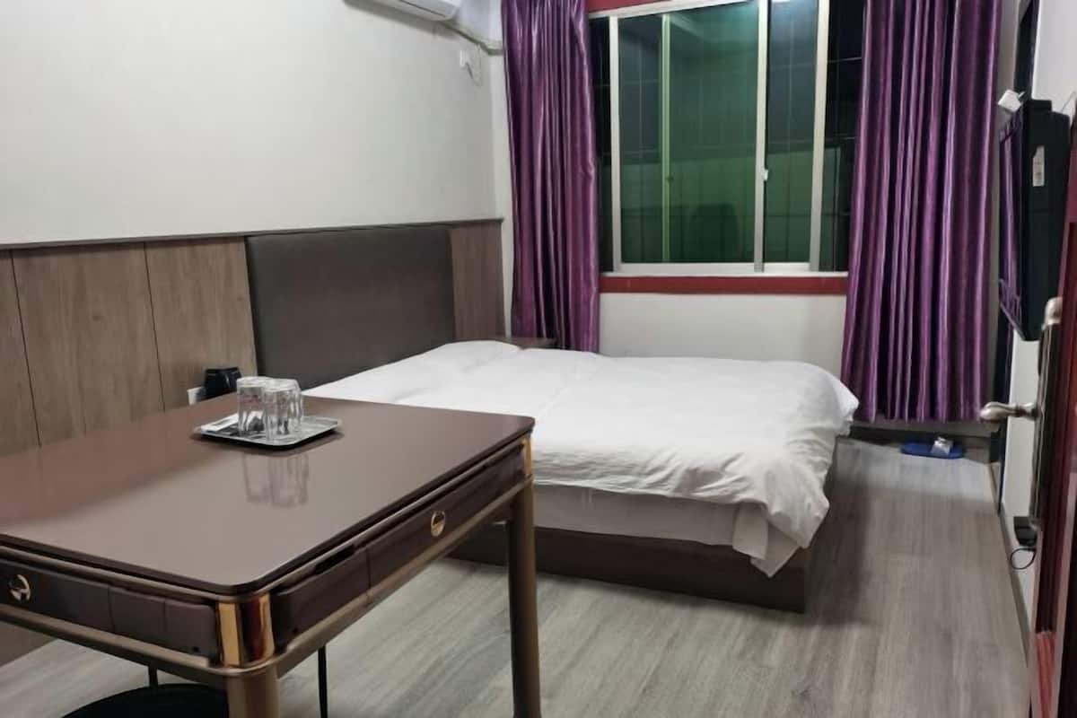 A hotel room with a bed, a wooden table with a glass, and purple curtains.
