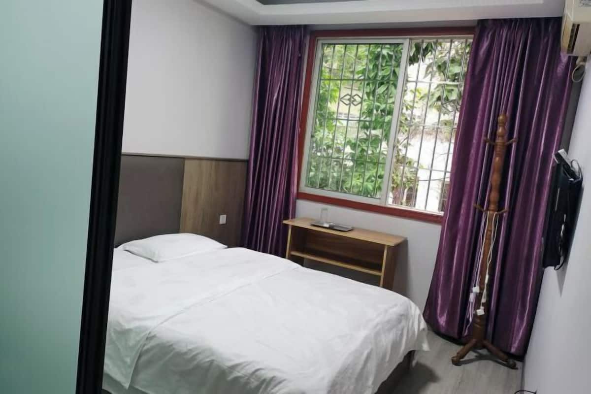 A bedroom with a bed, a wooden nightstand, a window with purple curtains, and a wooden wardrobe.
