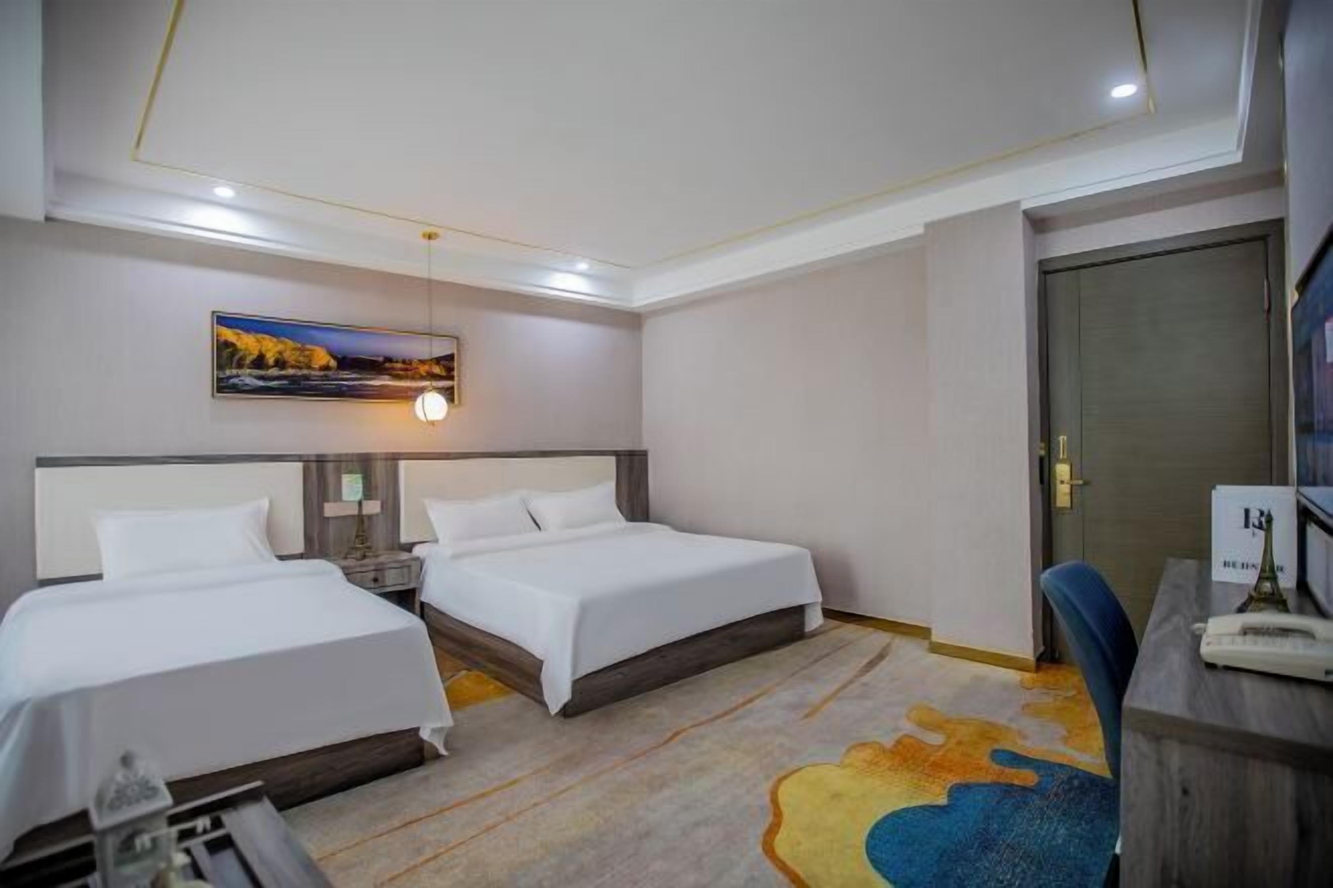 A hotel room with two beds, a desk, a chair, a desk lamp, a painting on the wall, and a door.