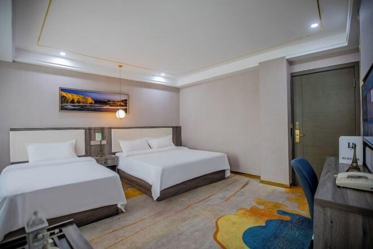 A hotel room with two beds, a desk, a chair, a desk lamp, a painting on the wall, and a door.