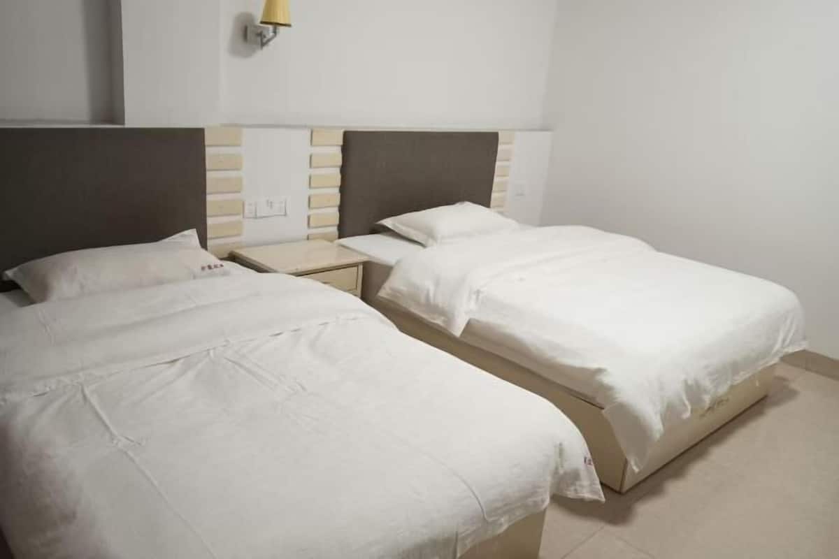 Twee enkele bedden met witte lakens, een nachtkastje, een wandlamp en een neutraal gekleurde muur.
