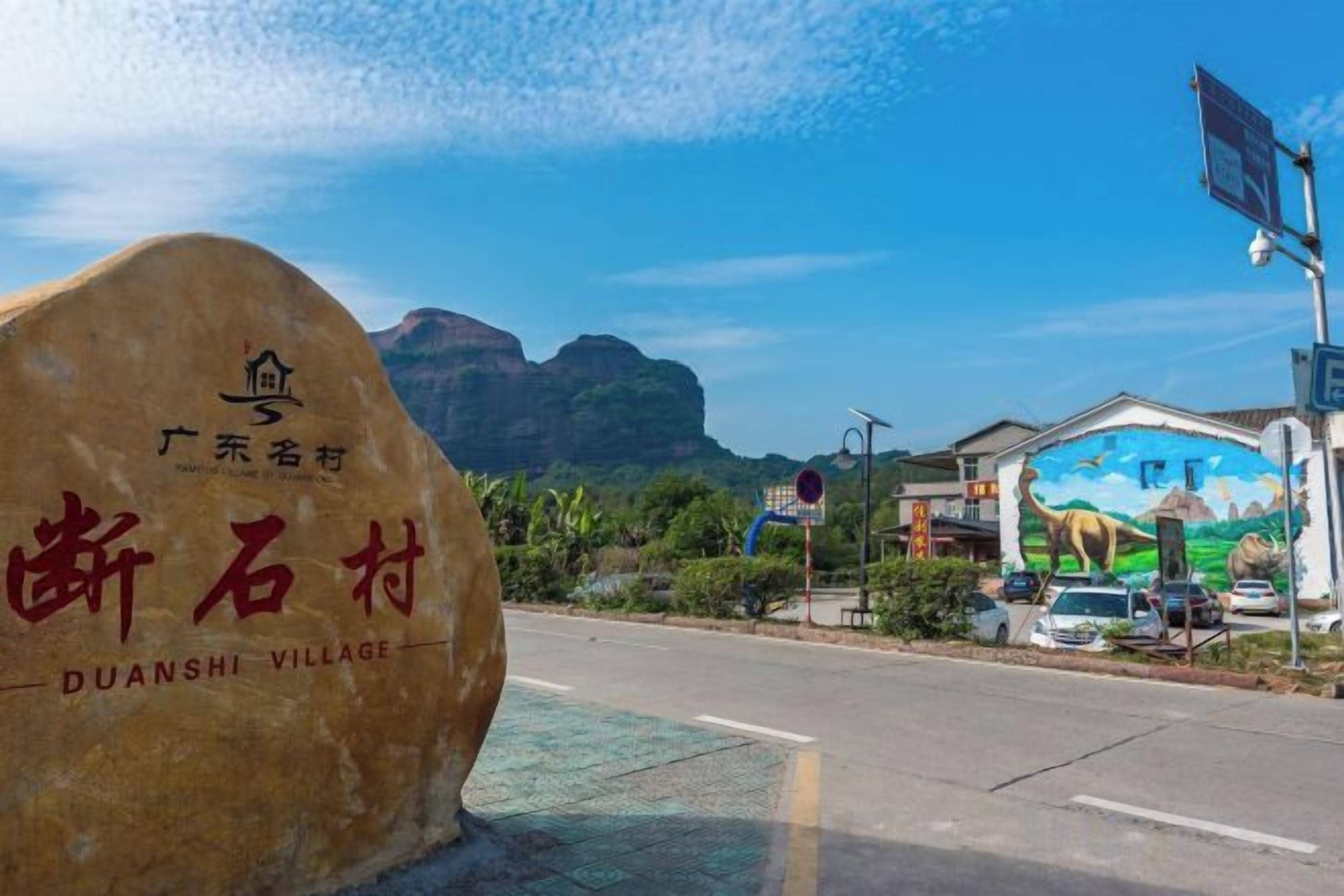 A large rock with 'GUANSHI VILLAGE' written on it, a mural on a building, and a road with cars.