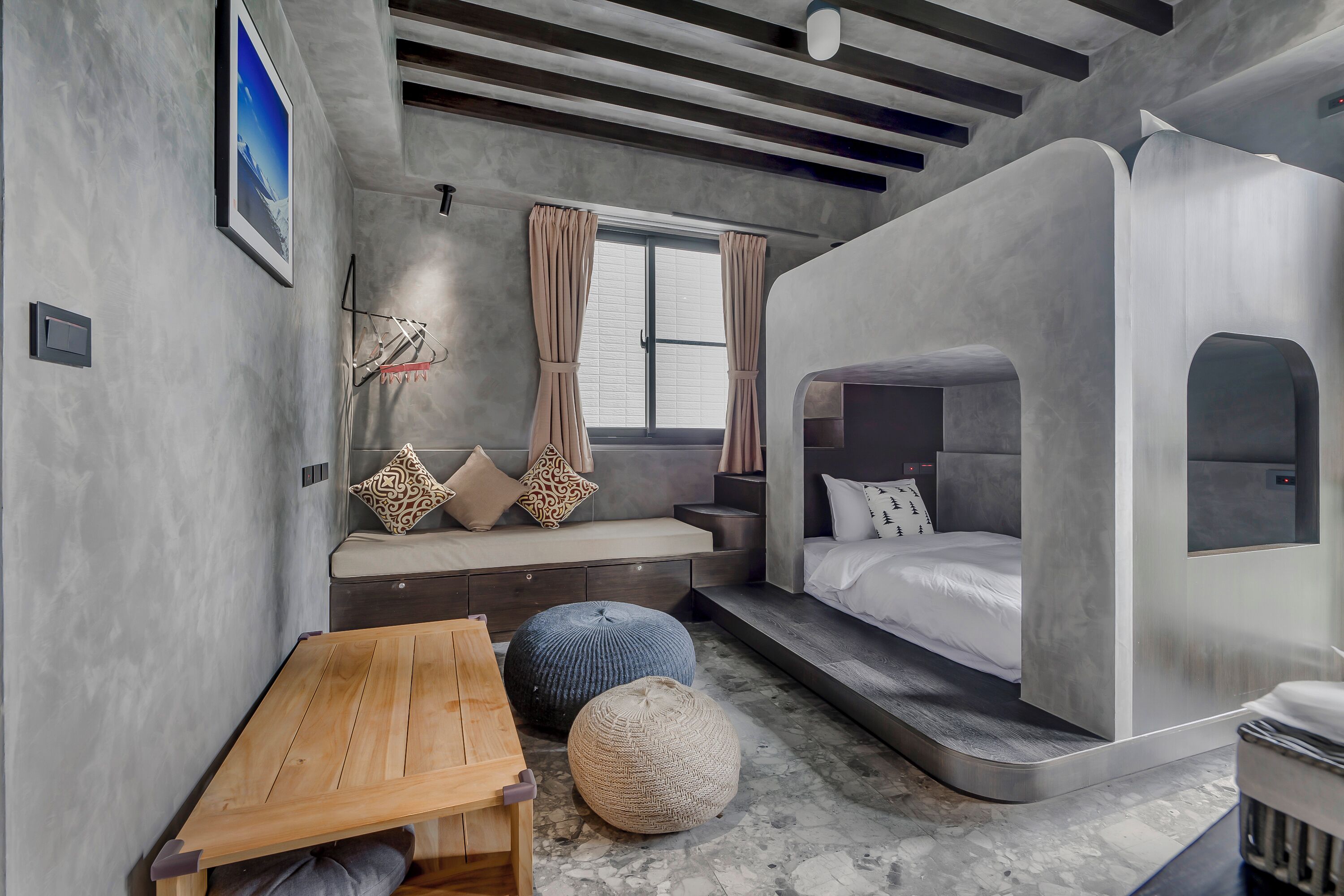 Un dormitorio moderno con cama empotrada, zona de estar con cojines, una mesa de madera y un otoman redonda.