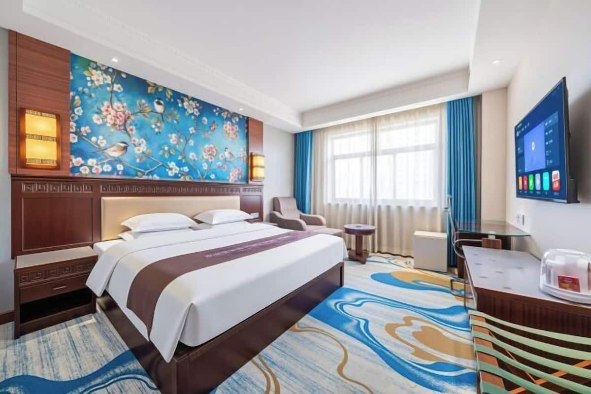 A hotel room with a bed, a desk, a chair, a TV, and a wall with a floral painting.