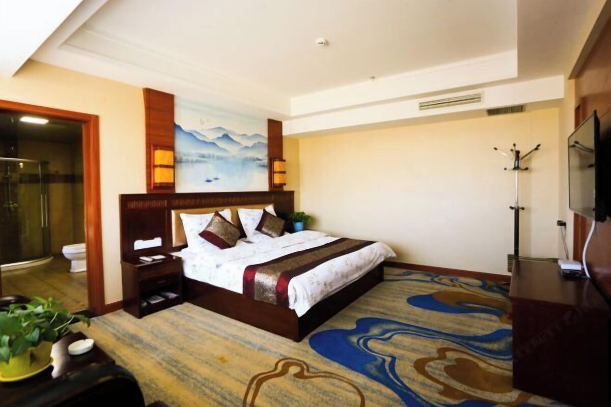 A hotel room with a bed, a shower, a toilet, a TV, and a painting on the wall.