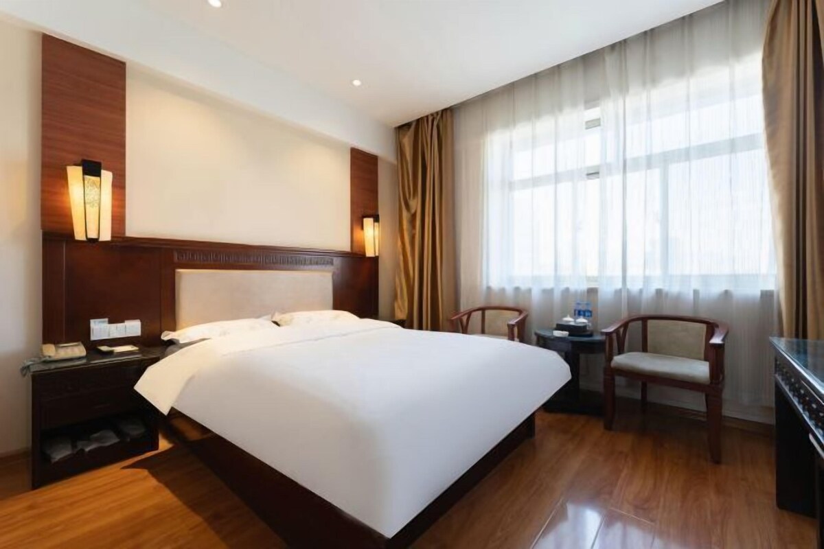 A hotel room with a large bed, a wooden nightstand, a desk with a chair, and a large window with curtains.