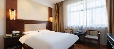 Habitación de hotel con una cama grande, una mesita de noche de madera, un escritorio con silla y un ventanal con cortinas.
