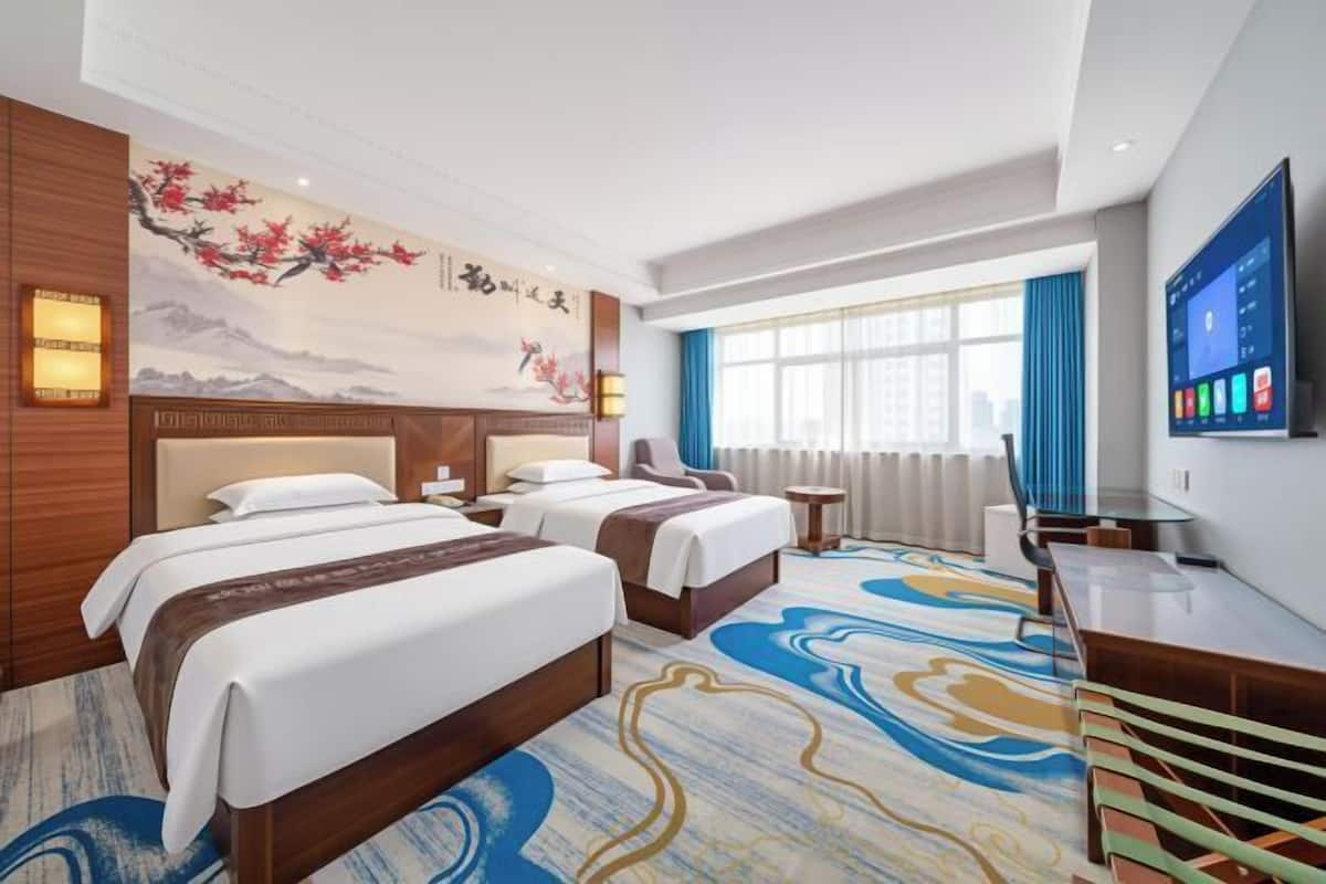 A hotel room with two beds, a desk, a TV, and a large window with curtains.
