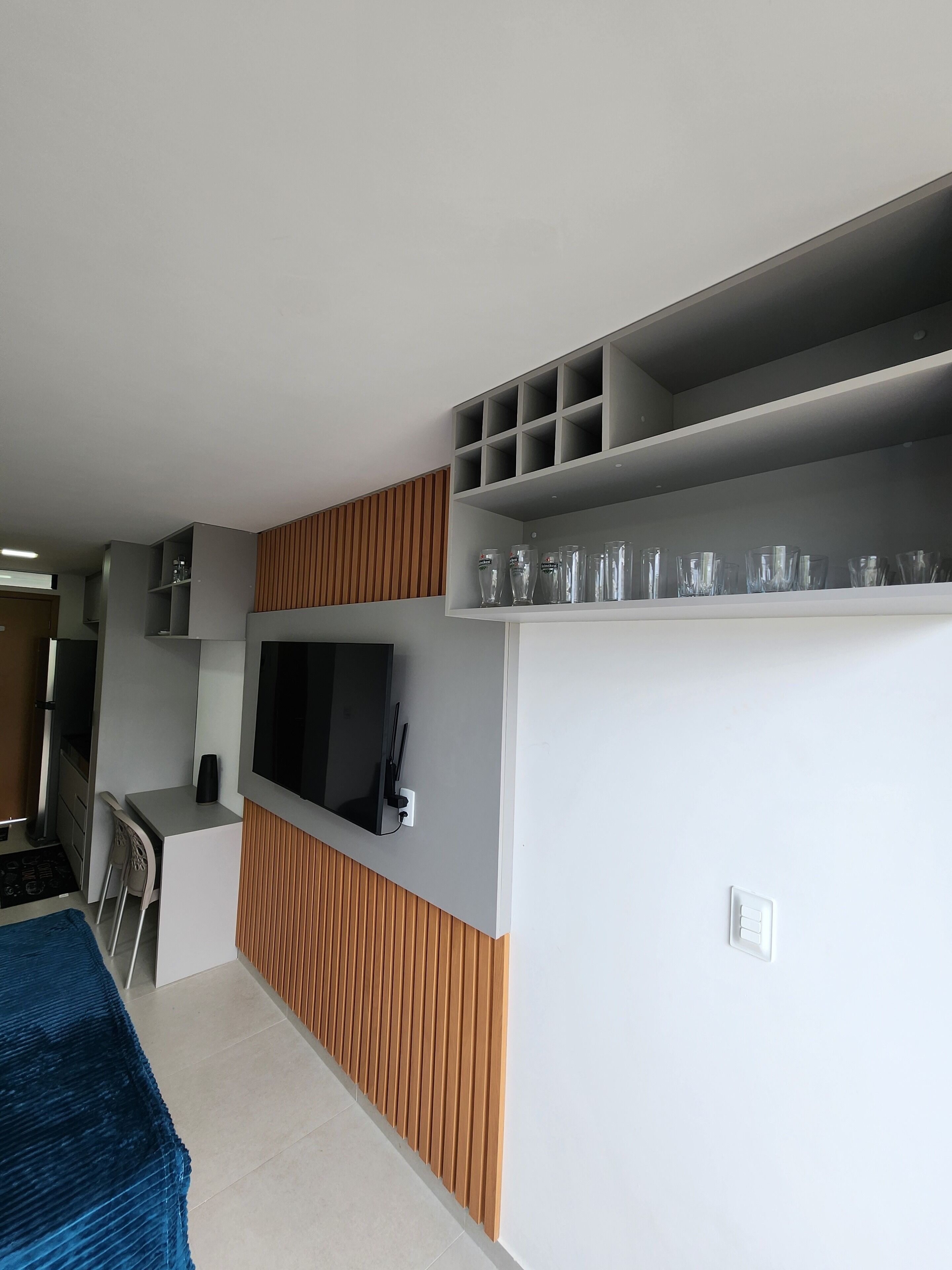 A modern hotel room with a flat-screen TV mounted on the wall, a small desk with a chair, and a built-in shelf with glasses.