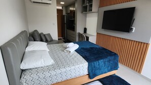 A modern hotel room with a bed, a desk, a TV mounted on the wall, and a kitchenette area. - Flat Ofurô 02 VP by La Playa (Ipojuca)