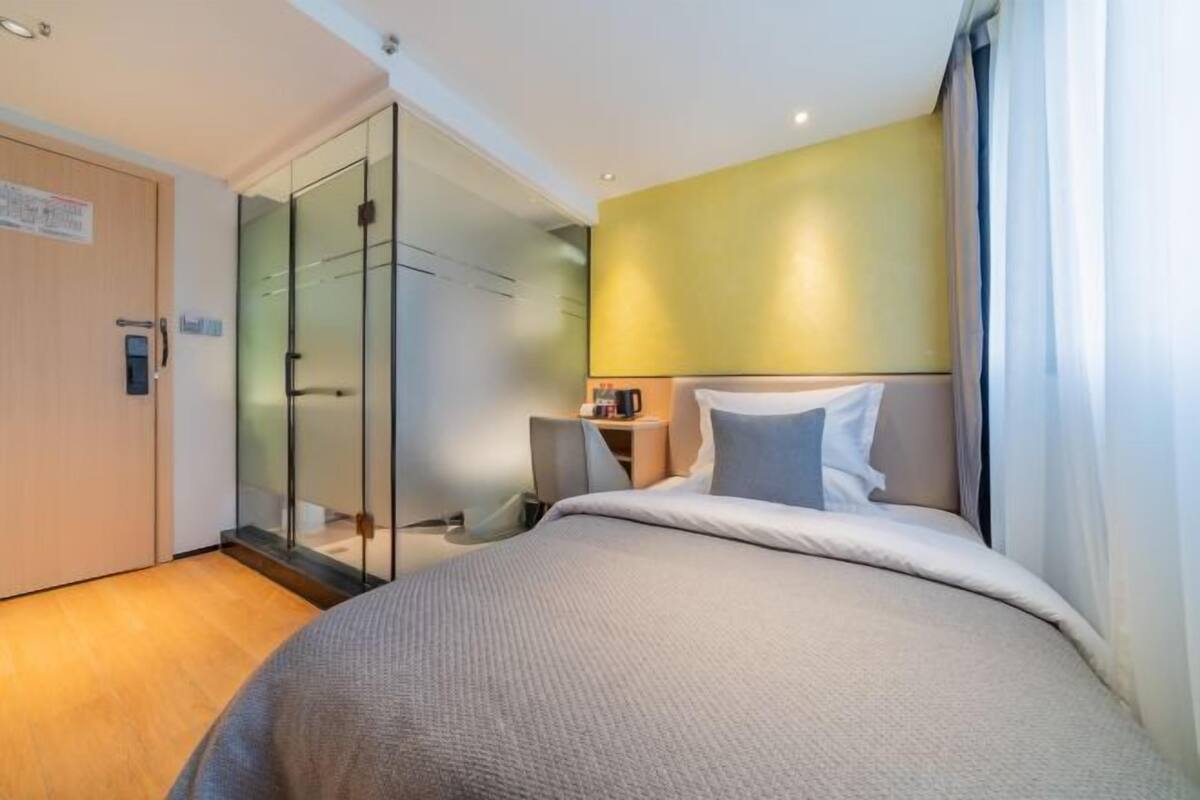 A hotel room with a bed, a bedside table, a nightstand, a glass partition, and a door.