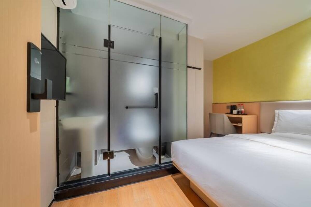 A hotel room with a glass partition, a bed, a nightstand, and a wall-mounted TV.