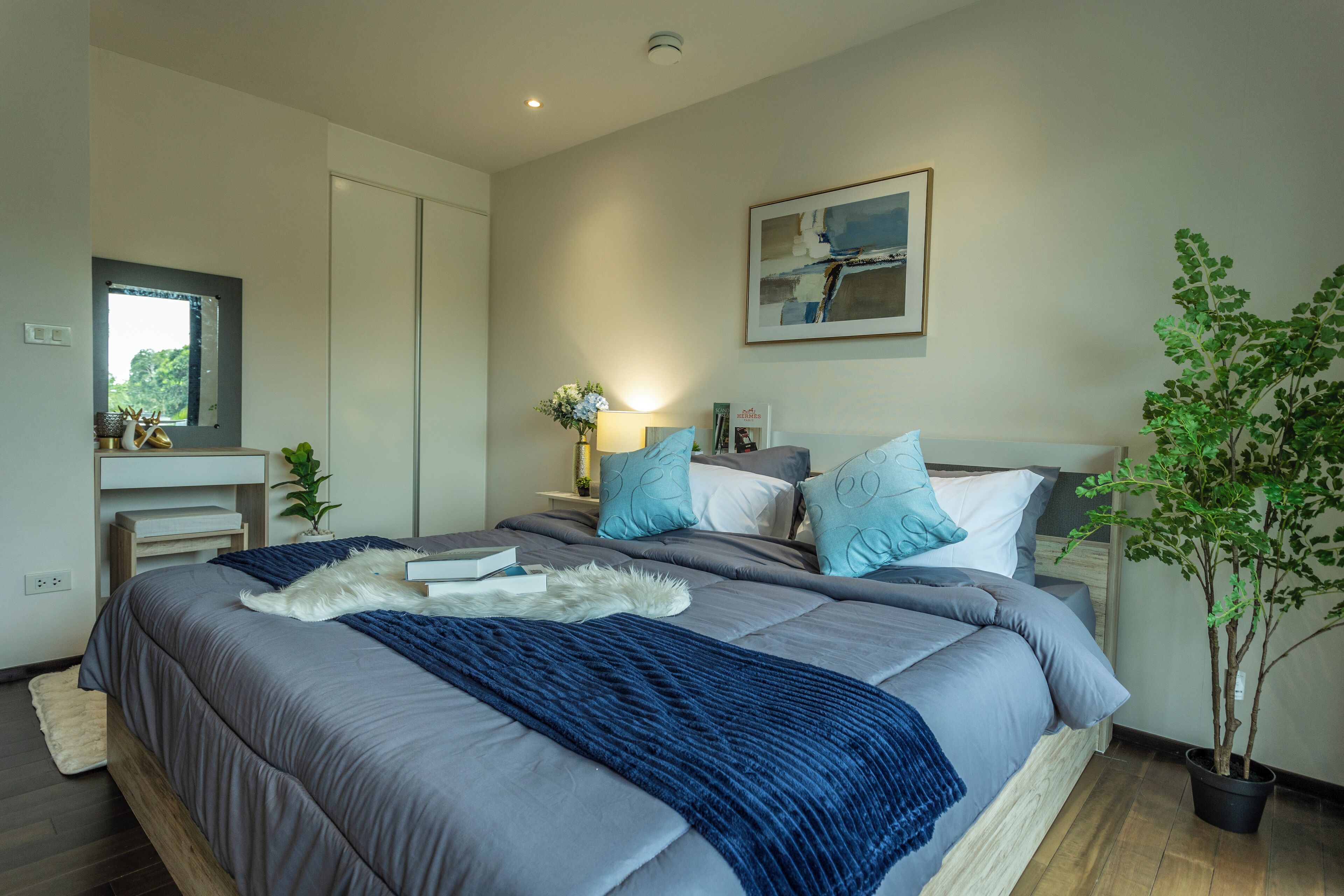 Un dormitorio moderno con una cama grande, una mesita de noche con una lámpara, una planta en maceta y una ventana con vistas a zonas verdes.