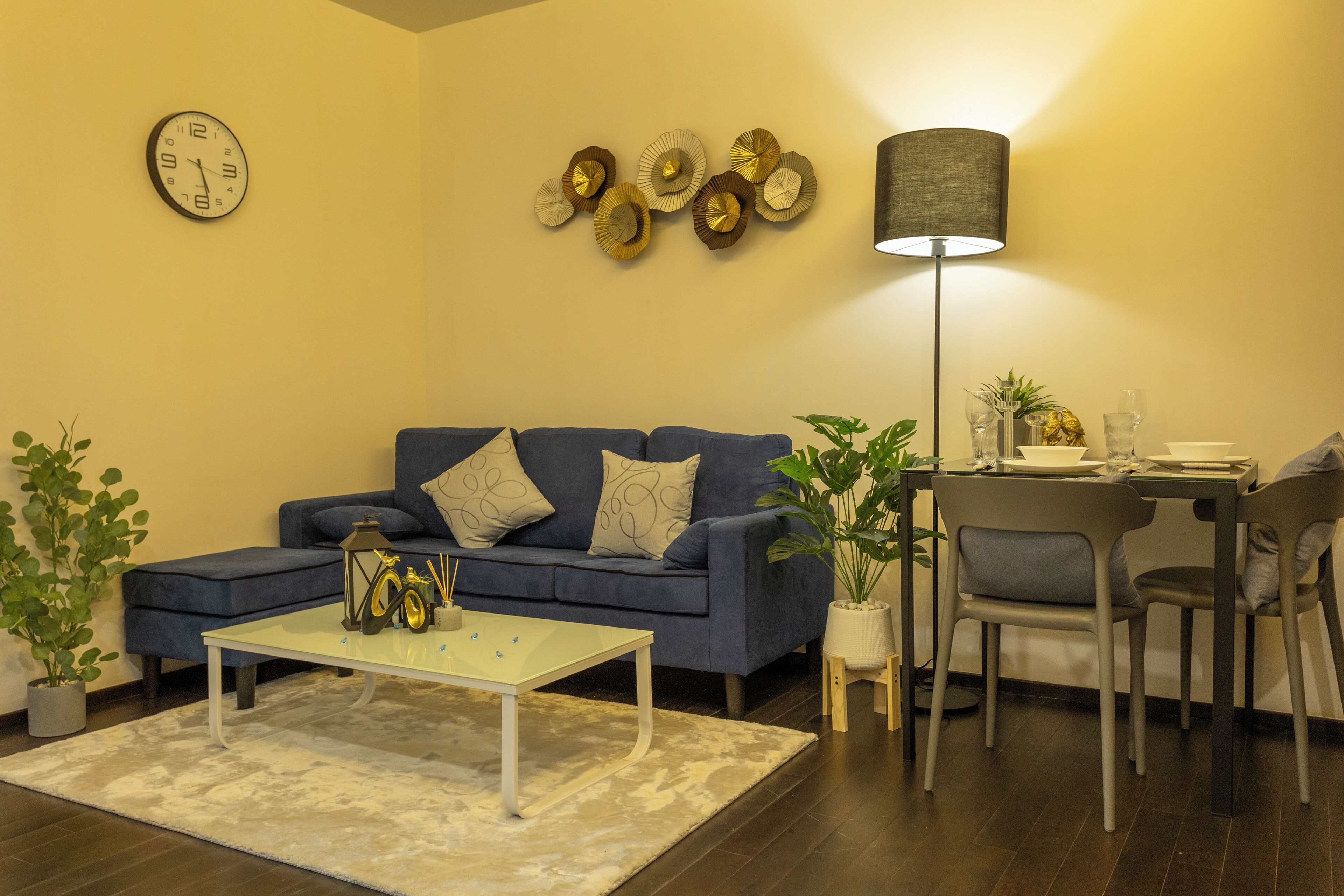 A living room with a blue sofa, a coffee table, a dining table with chairs, a lamp, and wall decorations.