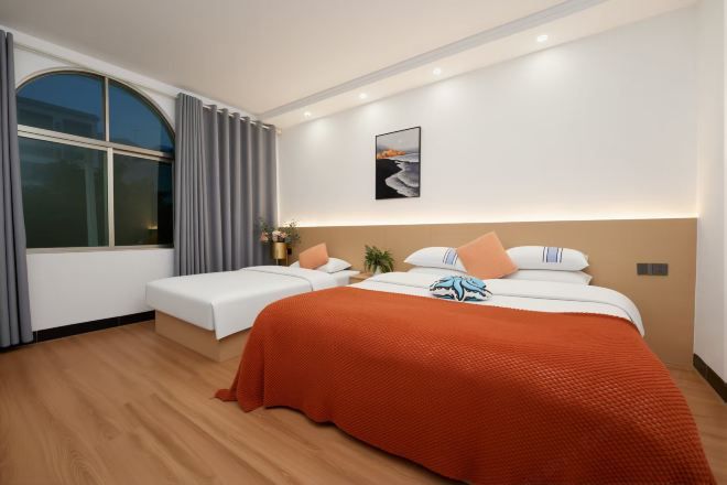 Quarto de hotel com uma cama grande, uma cama menor, uma janela com cortinas, uma obra de arte na parede e uma mesa de cabeceira com abajur.