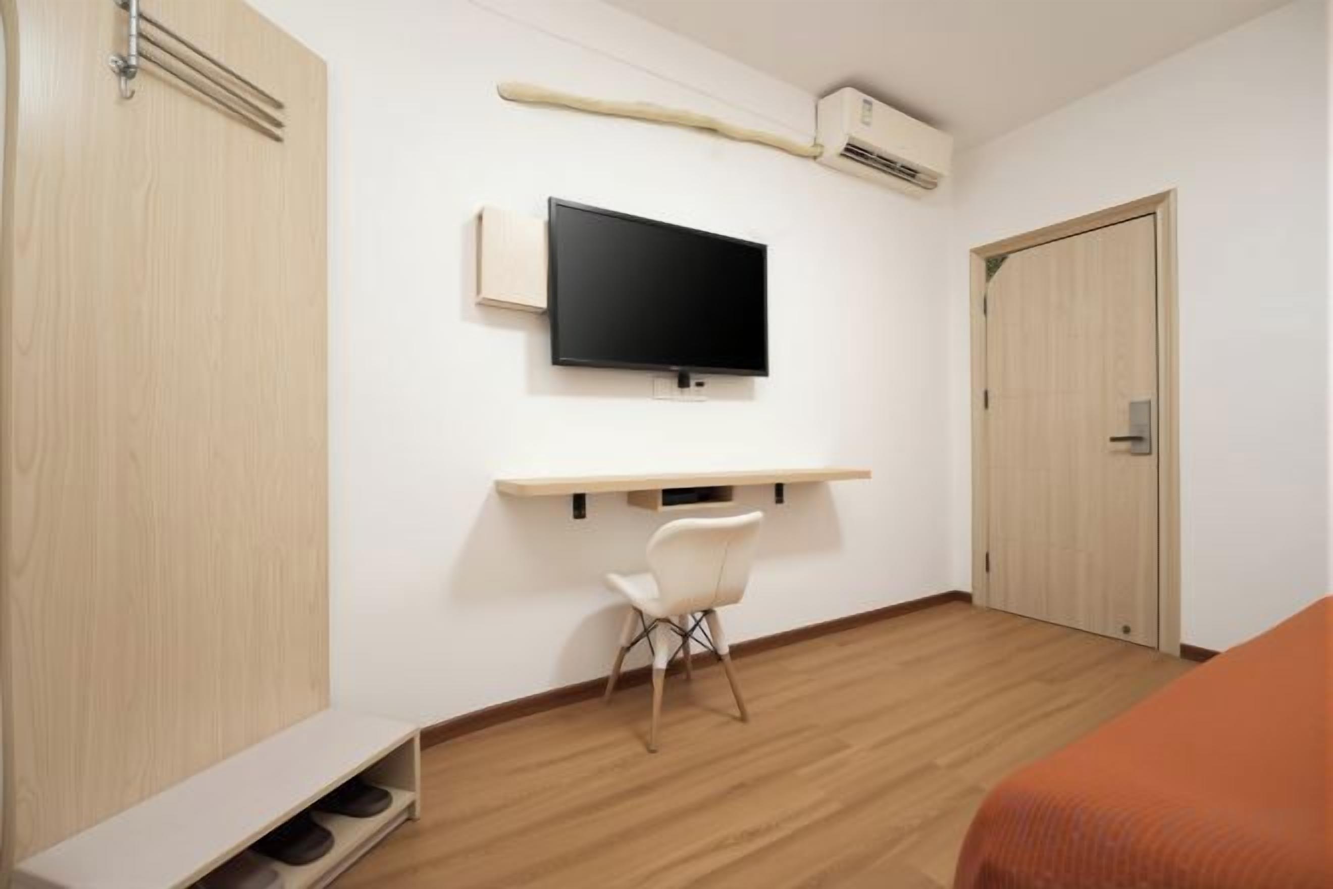A hotel room with a bed, a TV mounted on the wall, a desk with a chair, and a wardrobe.