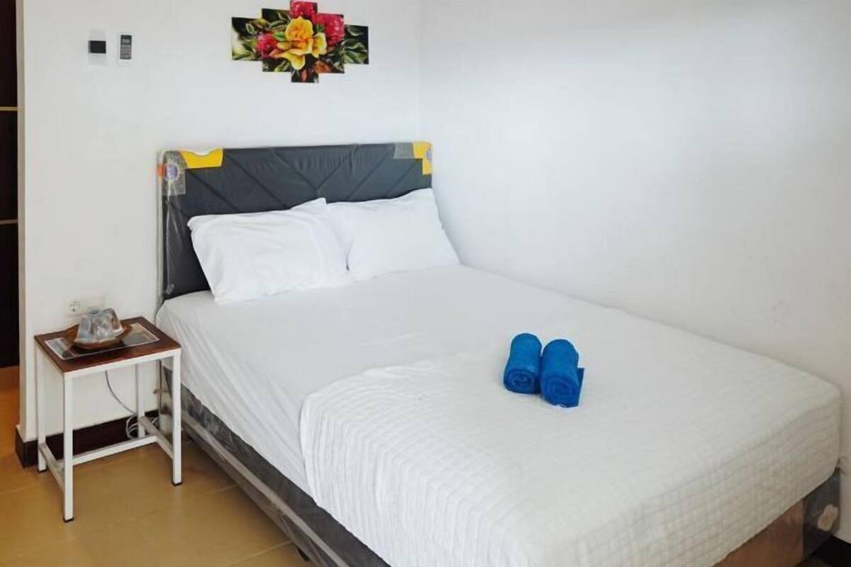 A neatly made bed with white bedding, a small bedside table with a kettle, and a wall decoration.