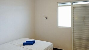 A single bed with a white bedspread and two blue pillows, a window with blinds, and a wall-mounted light switch. - RedDoorz Syariah near RSUD Mataram Lombok 2 (Mataram)