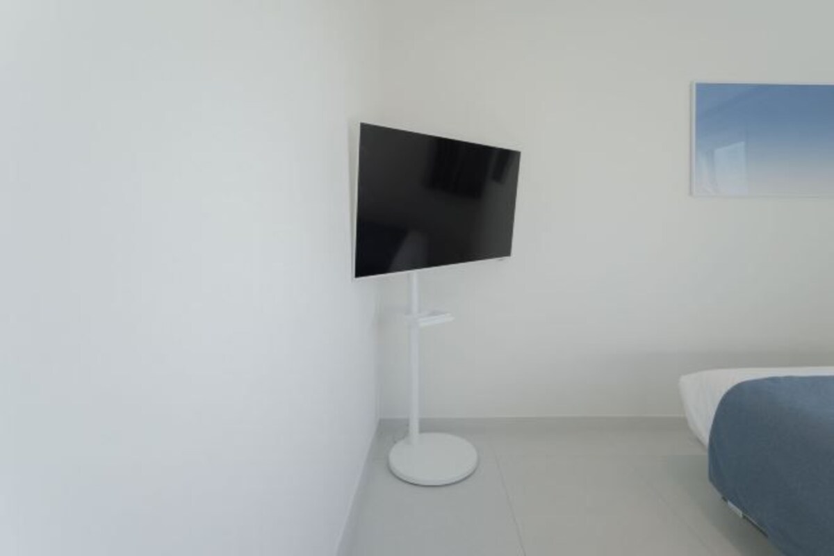 Una habitación de hotel moderna con un televisor de pantalla plana montado sobre un pedestal blanco, una cama con cubrecama azul y un cuadro en la pared.