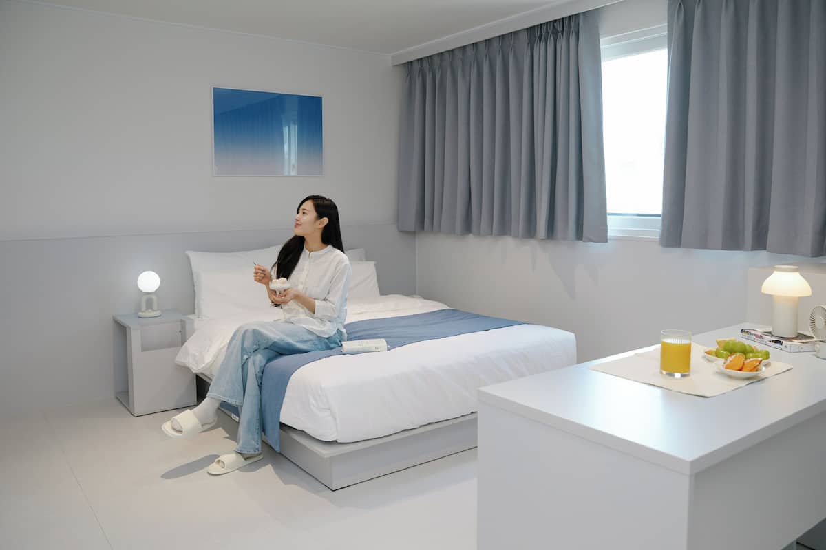 A modern hotel room with a bed, bedside table, and a desk with a lamp and fruit.