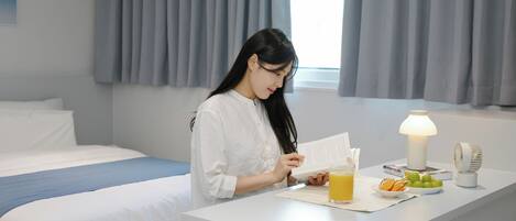 A hotel room with a bed, a bedside table with a lamp, a fan, and a breakfast tray with fruits and juice.