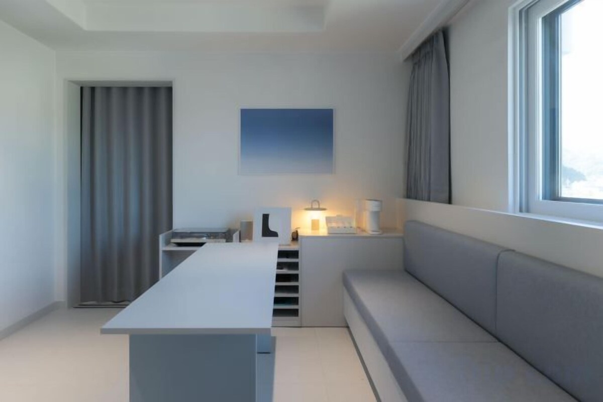 Una habitación moderna y minimalista con un escritorio largo y estrecho, un banco acolchado y un ventanal que proporciona luz natural.