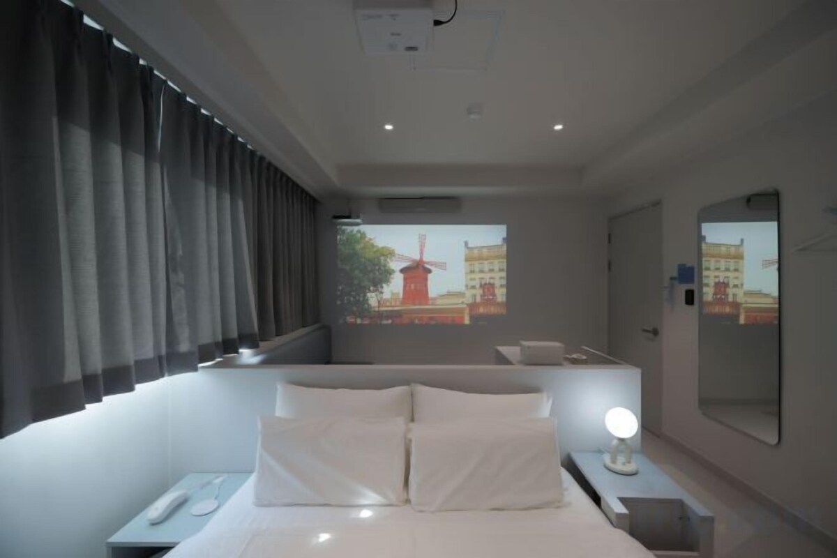 Habitación de hotel moderna con una cama grande, mesita de noche y vistas a un paisaje urbano a través del ventanal.