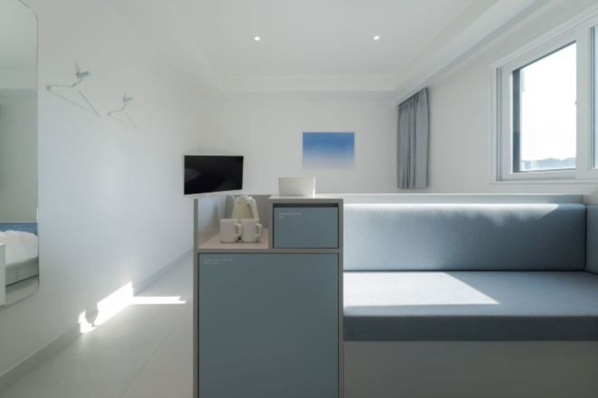 A modern hotel room with a minimalist design, featuring a light blue sofa, a small desk with a flat-screen TV, and a window providing natural light.