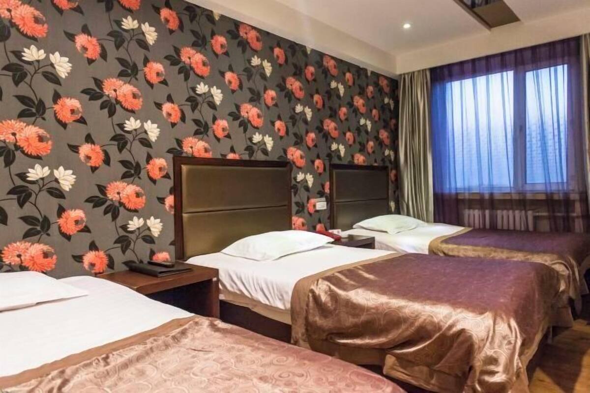 A hotel room with three beds, a floral wallpaper, and a large window with curtains.