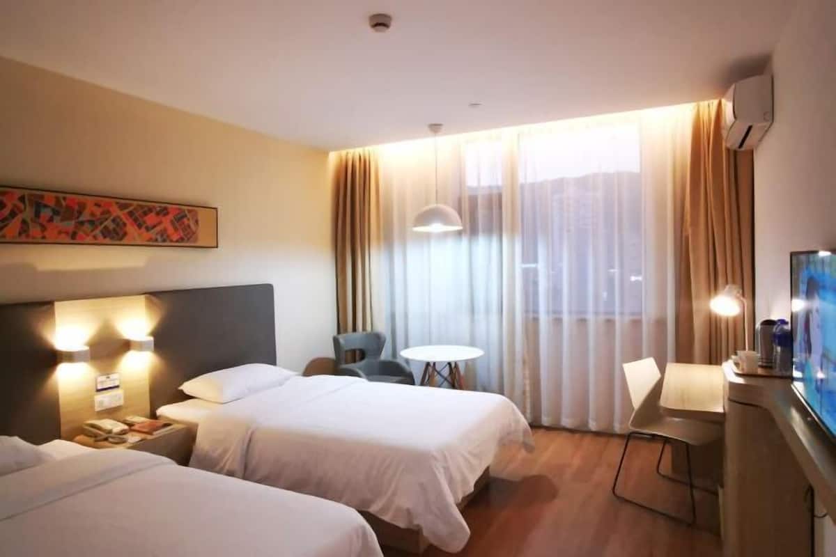 A hotel room with two beds, a desk, a chair, a small table, a TV, and a window with curtains.