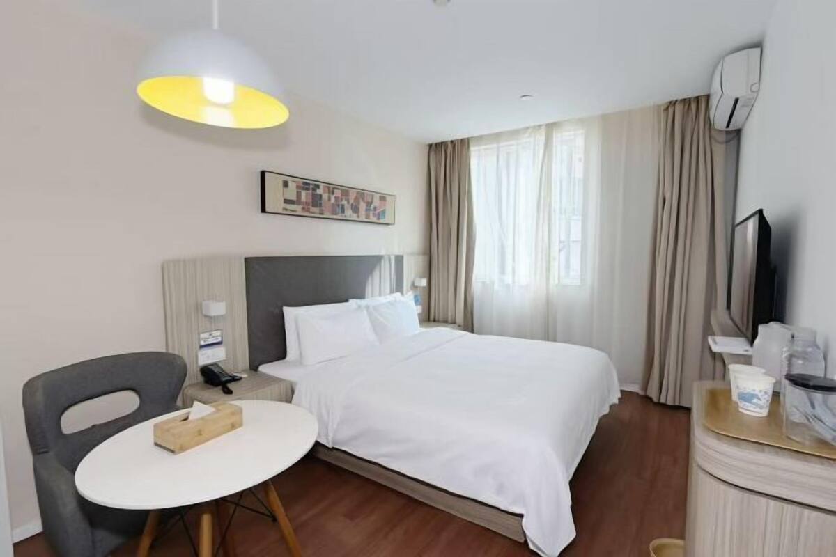A hotel room with a bed, a desk, a chair, a table, a lamp, a painting, an air conditioning unit, and a television.