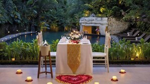 An outdoor dining area with a table set for two, surrounded by candles and rose petals, overlooking a pool and lush greenery. - Sakti Garden Resort & Spa (Ubud)