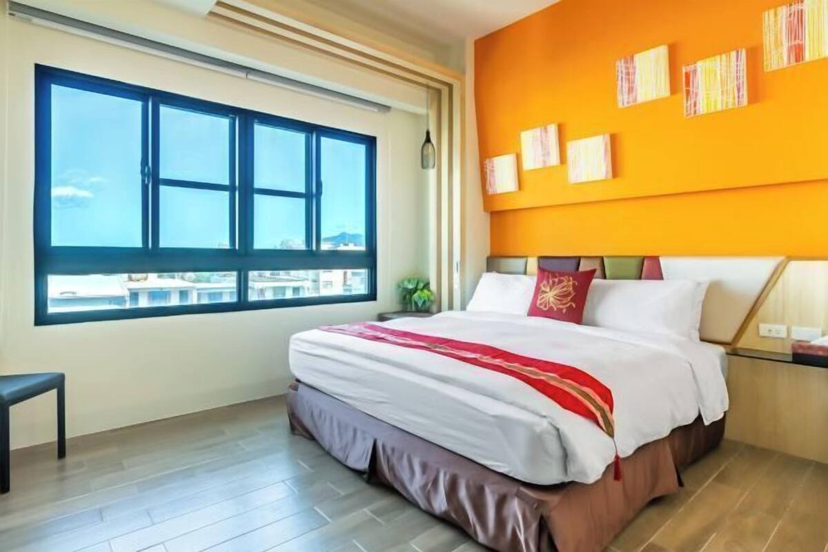 A hotel room with a large window, a bed with white bedding, a bench, and a wall with artwork.