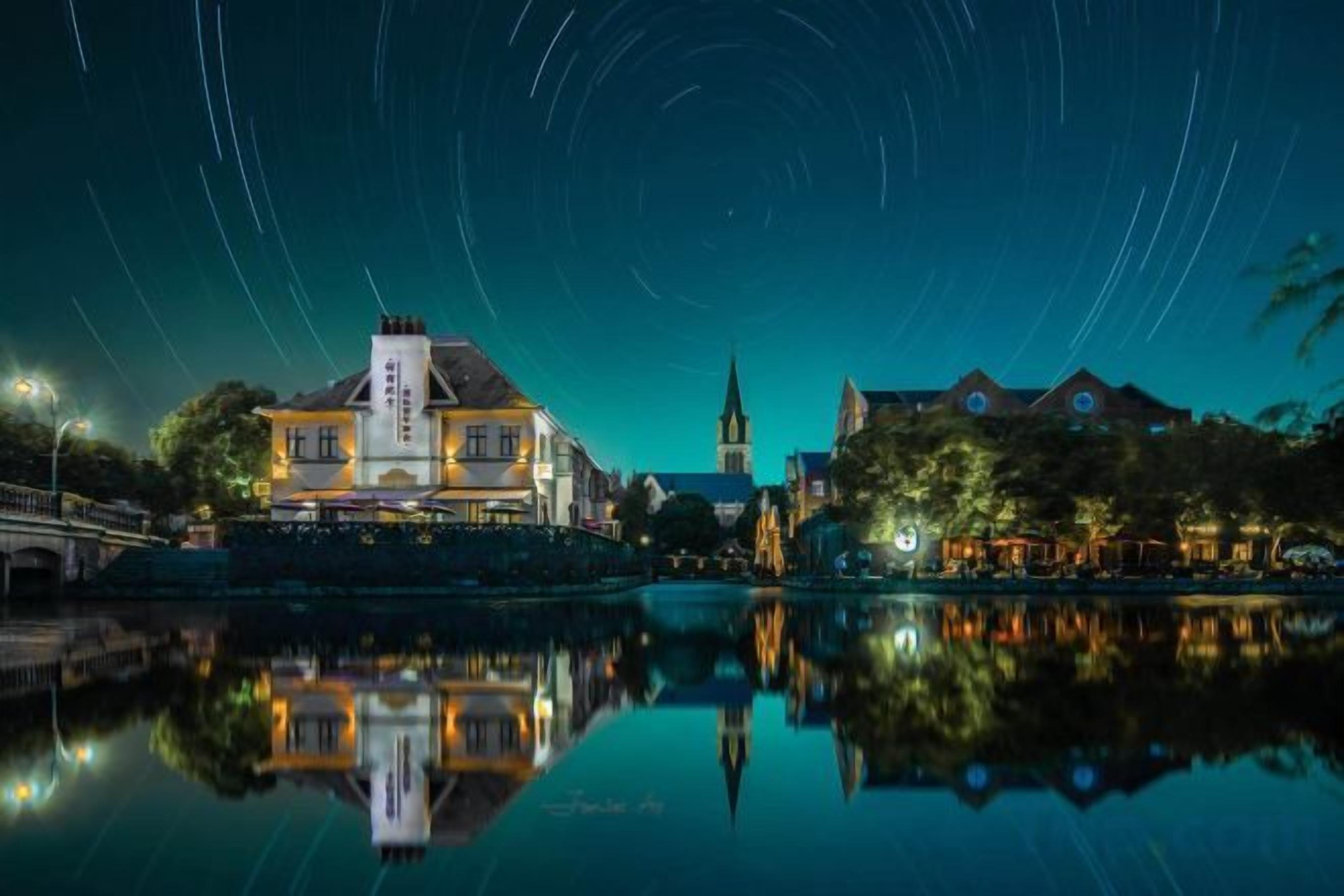 Un hotel frente al agua con su reflejo, una iglesia al fondo y un cielo nocturno despejado.