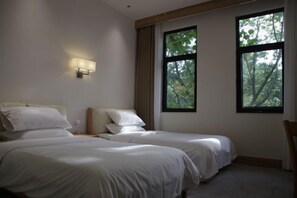 Habitación de hotel con dos camas individuales, una mesita de noche, una lámpara de pared y ventanas amplias con vistas a árboles.