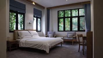 A bedroom with a large bed, two chairs, a nightstand, and a window with a view of greenery.