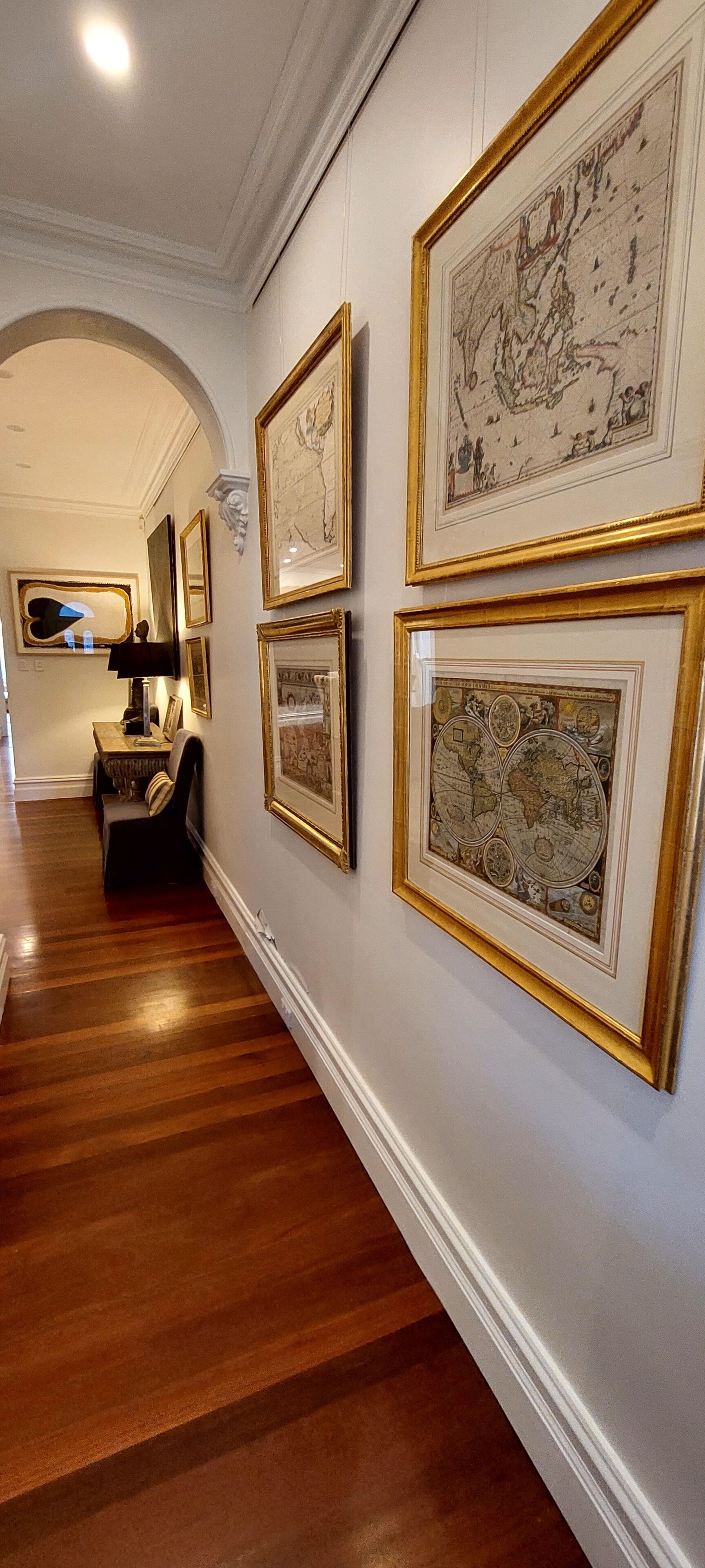 Un pasillo con suelo de madera, mapas enmarcados en la pared y una mesa con lámpara.