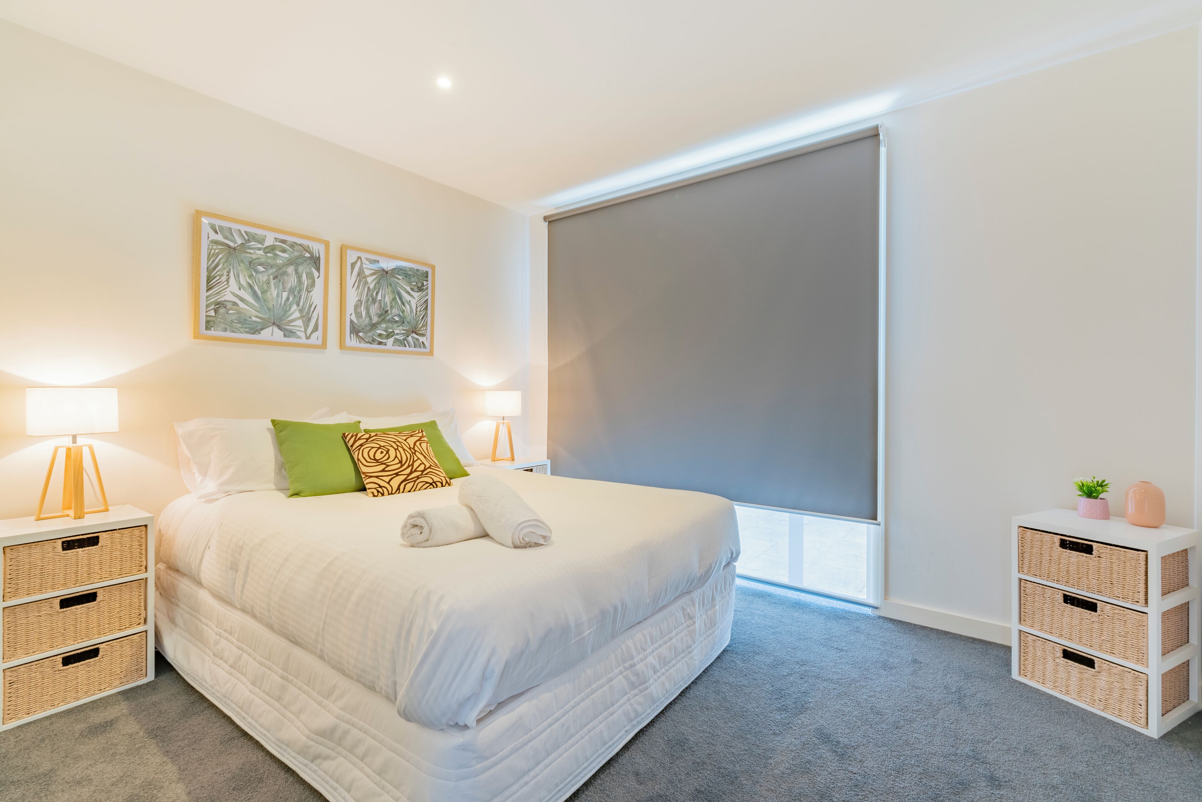 Um quarto moderno com uma cama grande, duas mesas de cabeceira com luminárias, uma janela ampla com persiana e duas gravuras botânicas emolduradas na parede.