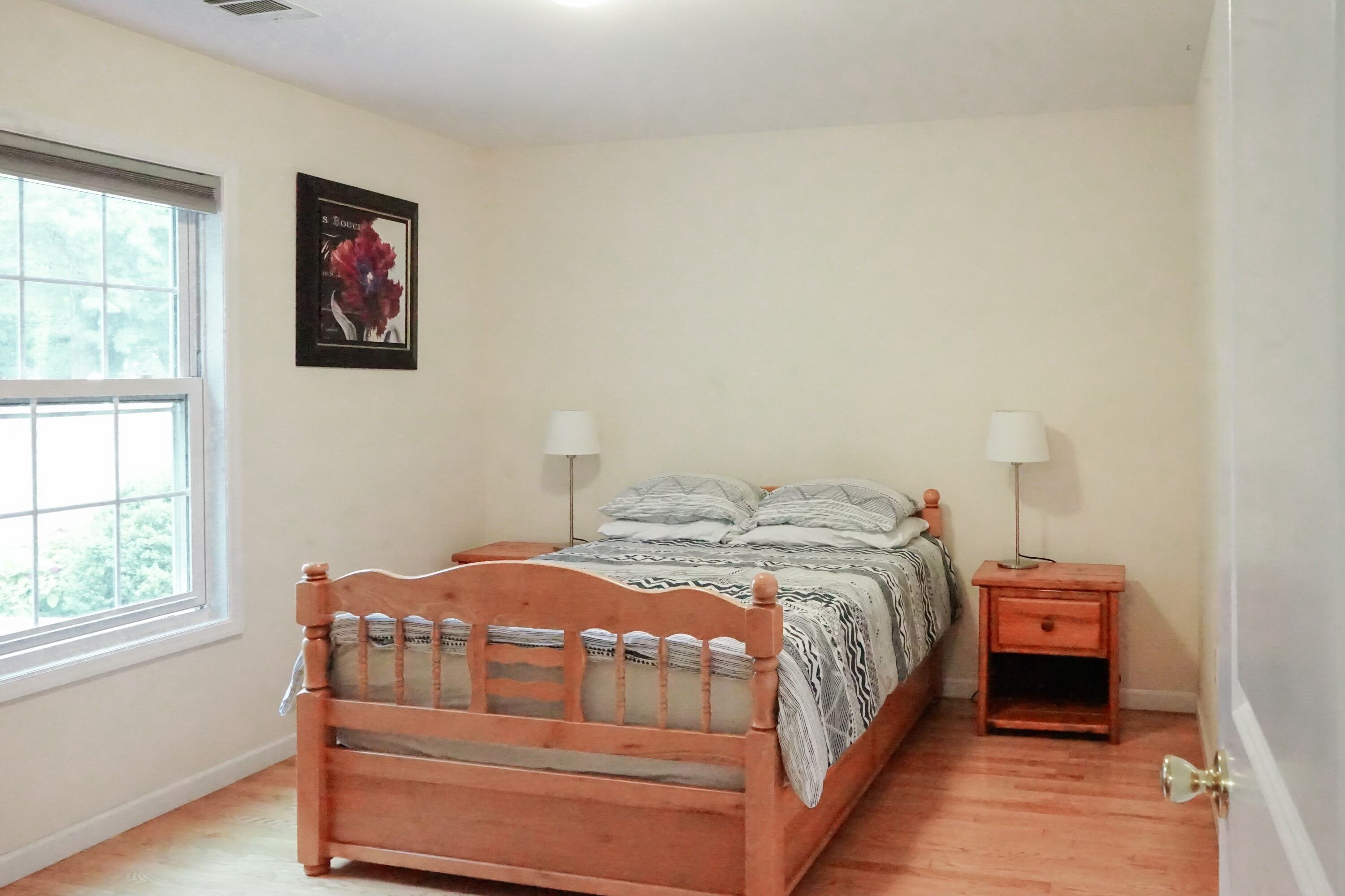 A bedroom with a wooden bed, two bedside tables with lamps, a window, and a framed picture on the wall.