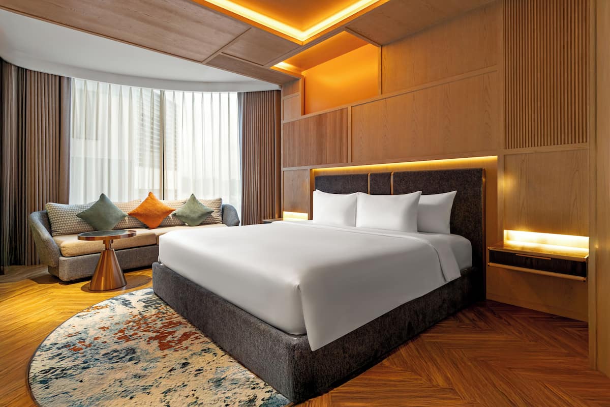 Ein modernes Hotelzimmer mit einem großen Bett, einer Couch mit Kissen, einem kleinen Beistelltisch und einem gemusterten Teppich.